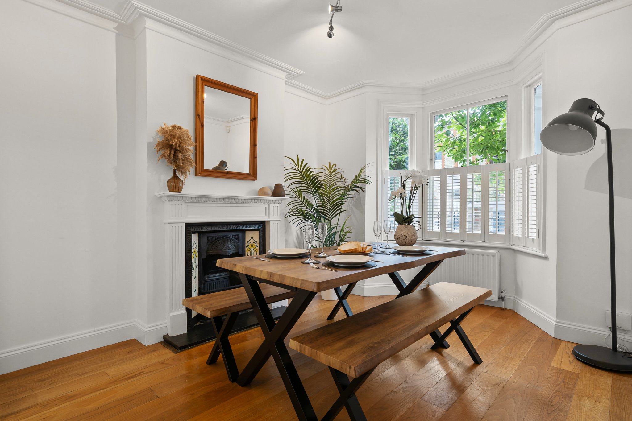 A dining room with a wooden table and benches, white walls, a fireplace with decorative tiles, a mirror above the fireplace, a window with shutters, potted plants, and a standing floor lamp.