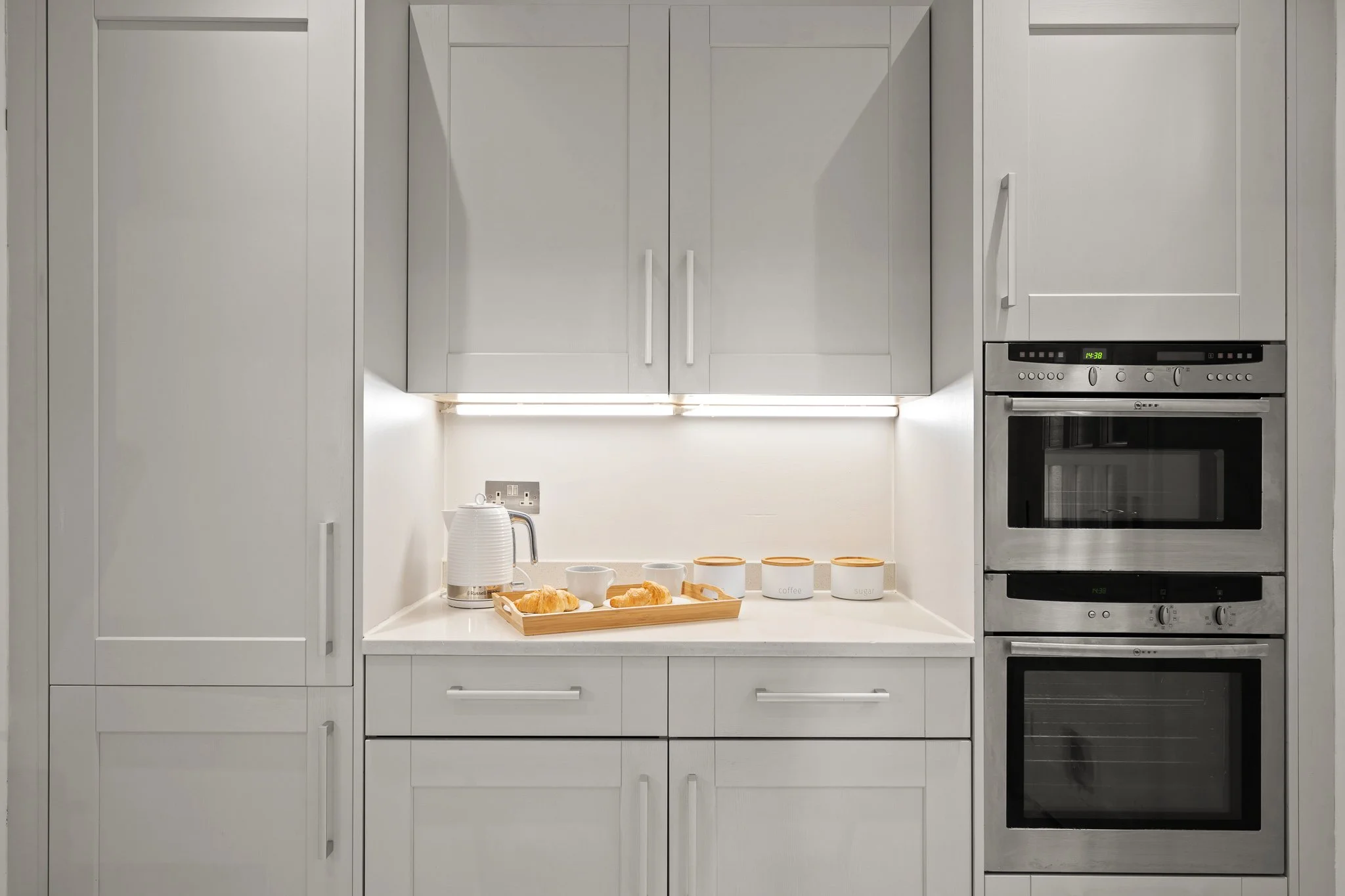 White kitchen with built-in appliances, table with croissants, cups, and jars for sugar and coffee, under overhead cabinets, with under-cabinet lighting.