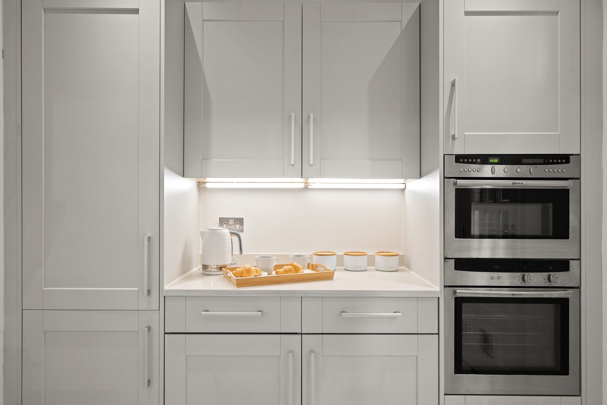 White kitchen with built-in appliances, table with croissants, cups, and jars for sugar and coffee, under overhead cabinets, with under-cabinet lighting.
