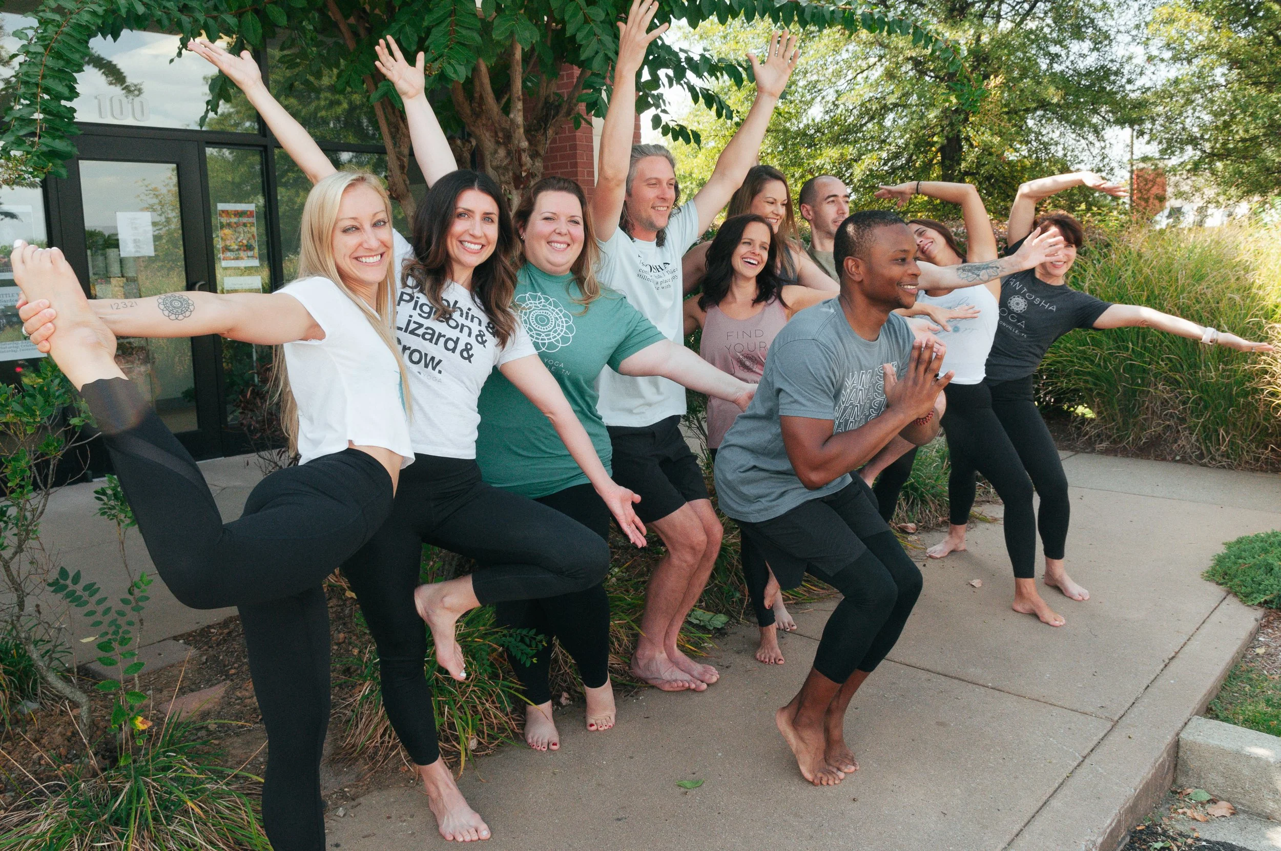Group of people doing yoga poses outside on a sidewalk, with trees and bushes in the background.