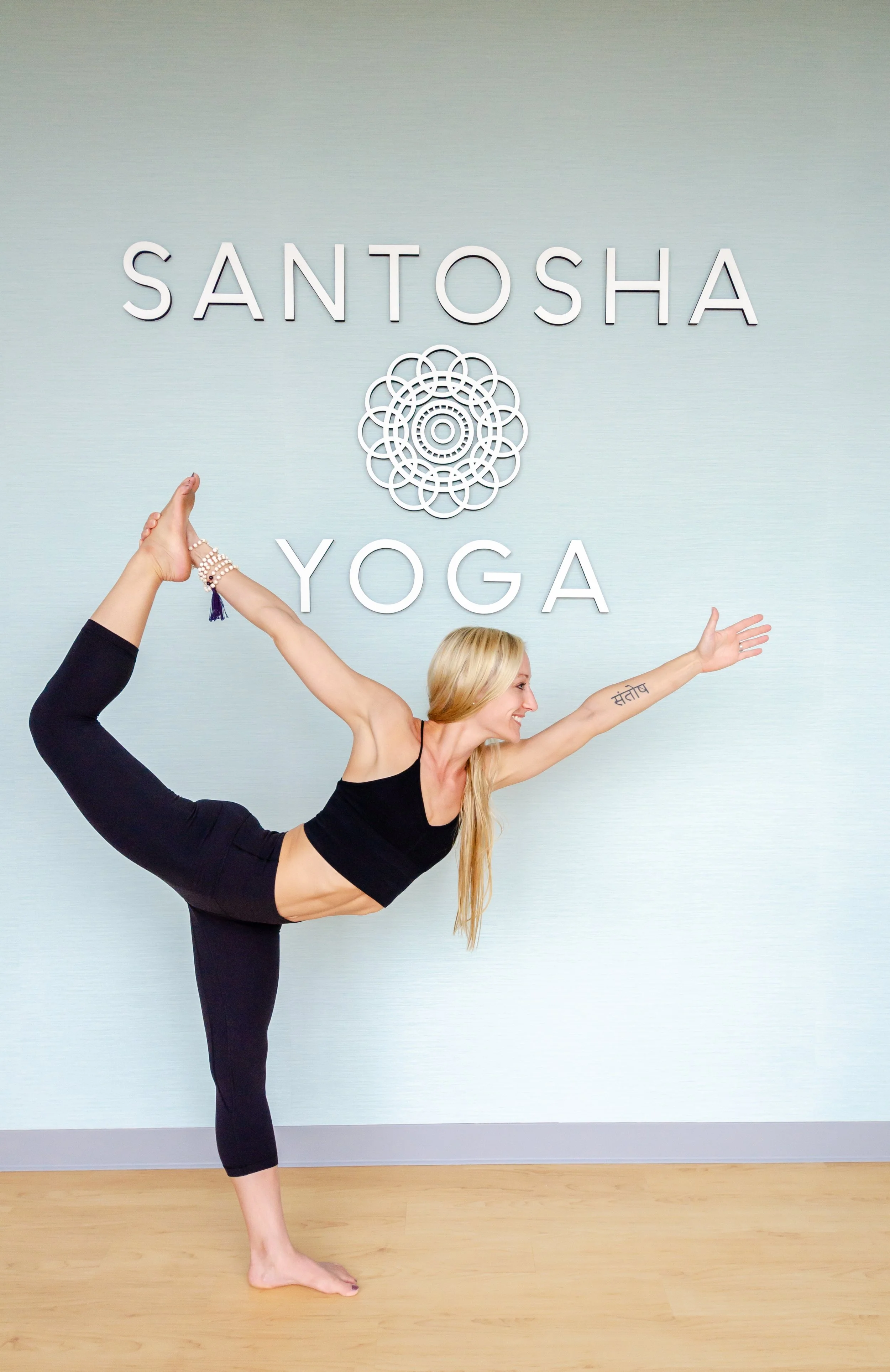 A woman practicing yoga in front of a wall with the sign 'SANTOSHA YOGA.' She is in a standing bow pose, balancing on one leg with the other leg bent backward and holding her foot with one hand, reaching forward with the other arm.