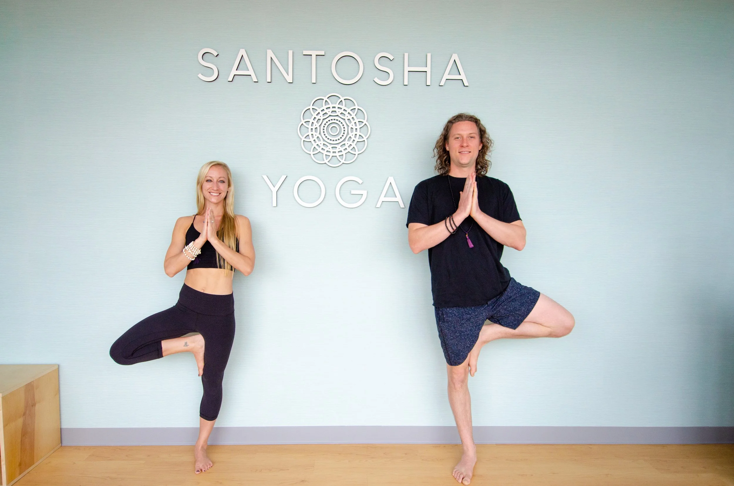 Two people practicing yoga in front of a sign that says 'SANTOCHA YOGA' on a light blue wall. The woman on the left is standing on her right leg with her left foot resting on her right inner thigh, hands in prayer position. The man on the right is balancing on his left leg with his right foot resting on his left inner thigh, hands in prayer position. Both are smiling.