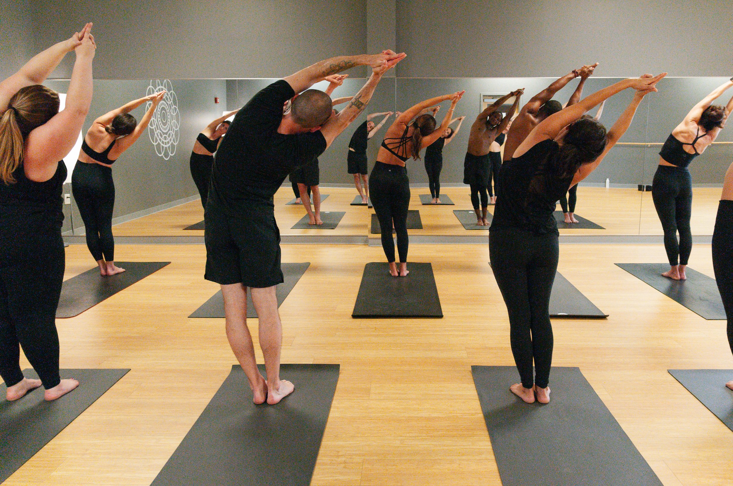 Group of people practicing yoga in a studio, all in a forward bend pose with arms extended overhead, standing on black yoga mats on a wooden floor.