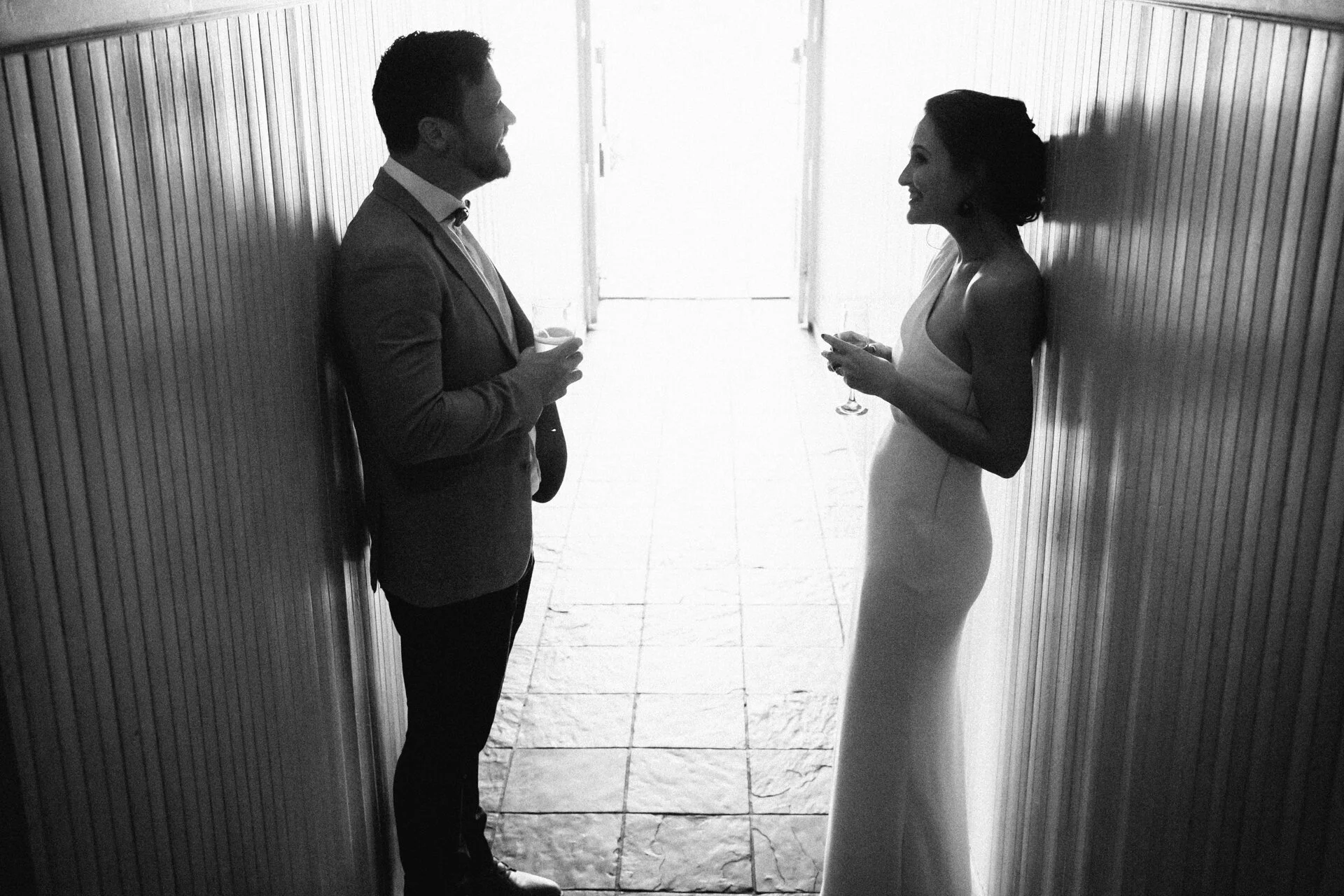 A black-and-white photo of a man and woman standing in a narrow hallway, facing each other, both holding glasses of what appears to be champagne, smiling and engaging in conversation.