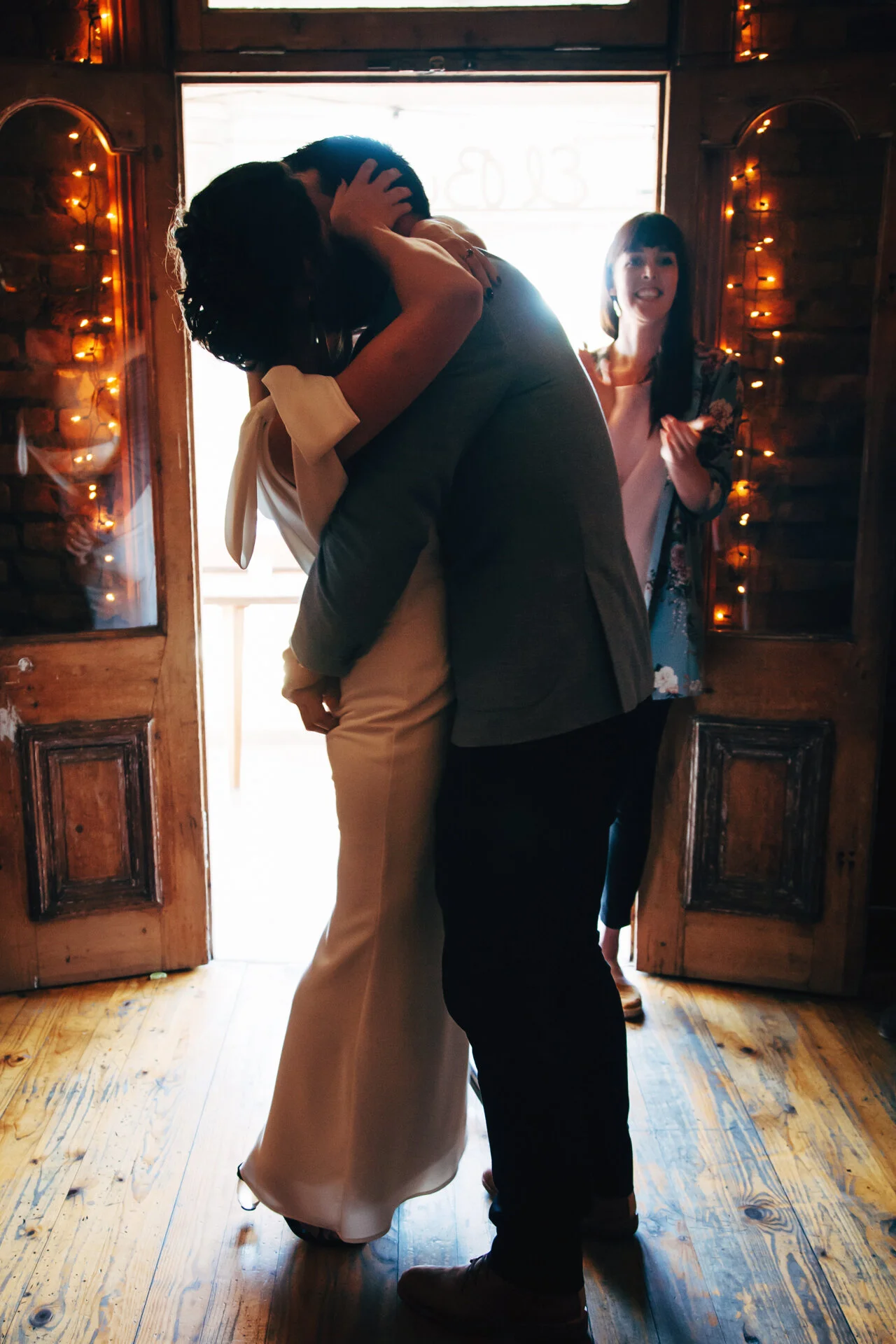 A couple sharing a kiss inside a warmly lit room with wooden walls and fairy lights, while a woman in the background cheers.
