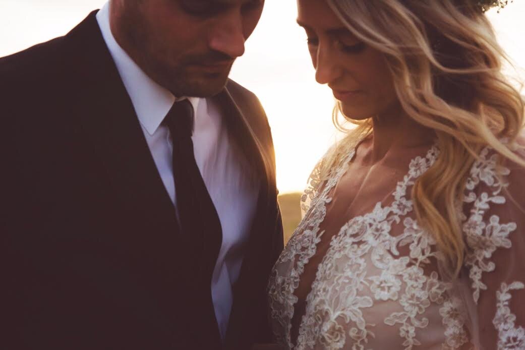 A bride and groom stand close together outdoors at sunset, both with heads bowed, serious expressions, and eyes closed. The woman wears a lace dress and has long wavy hair, while the man wears a suit and tie.