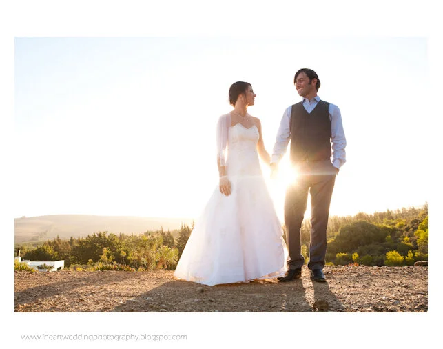 Couple in wedding attire holding hands outdoors at sunset with a landscape in the background.