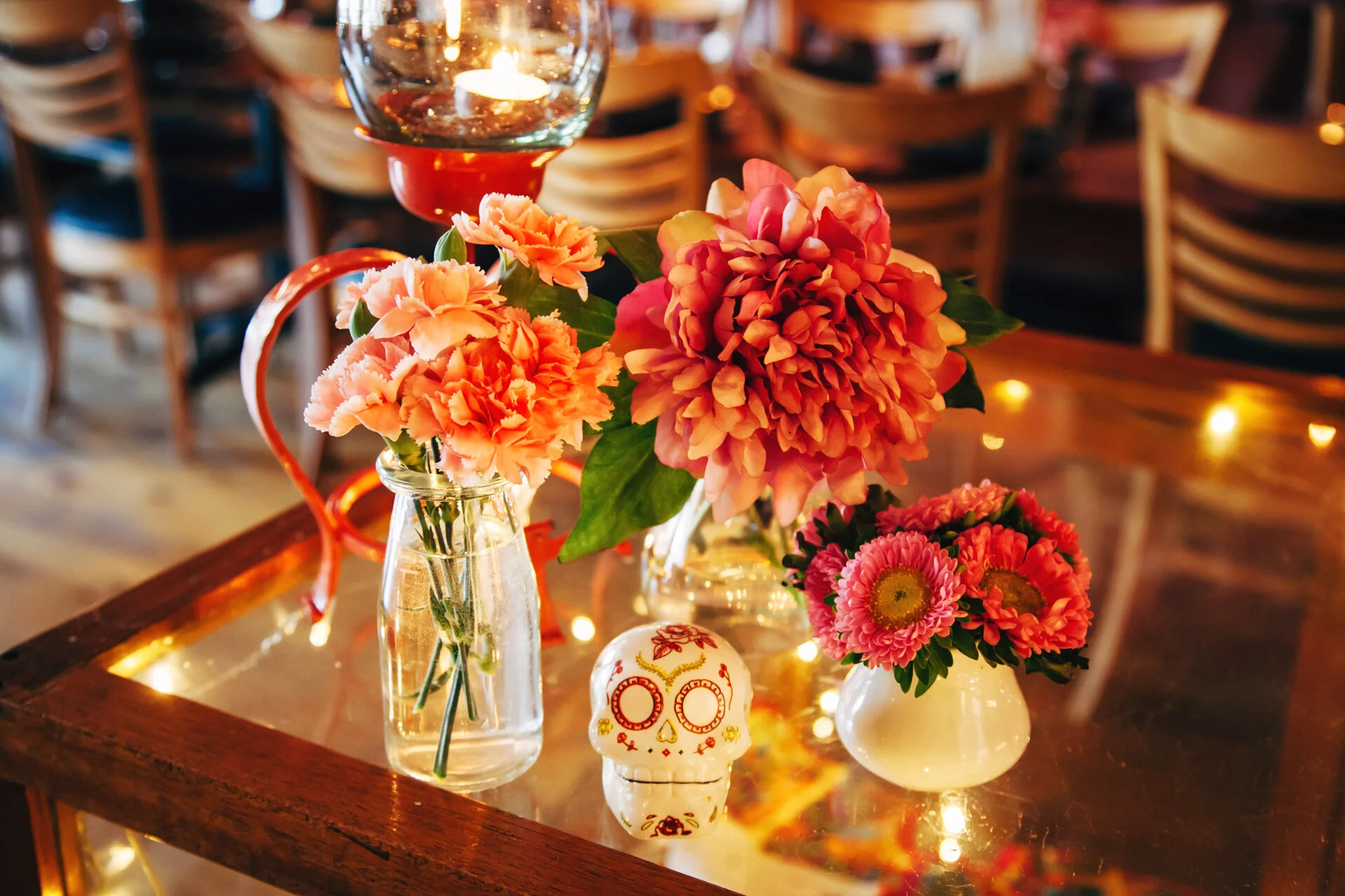 Vases with pink and orange flowers on a wooden table, decorated for celebration, with a sugar skull ornament and a wine glass with a lit candle inside.