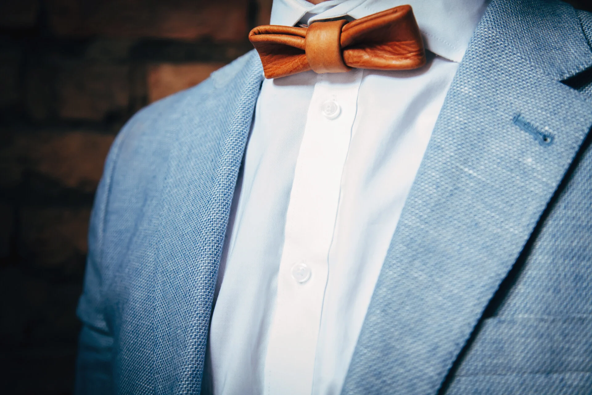 Close-up of a man wearing a light blue textured blazer, white dress shirt, and a brown leather bow tie.