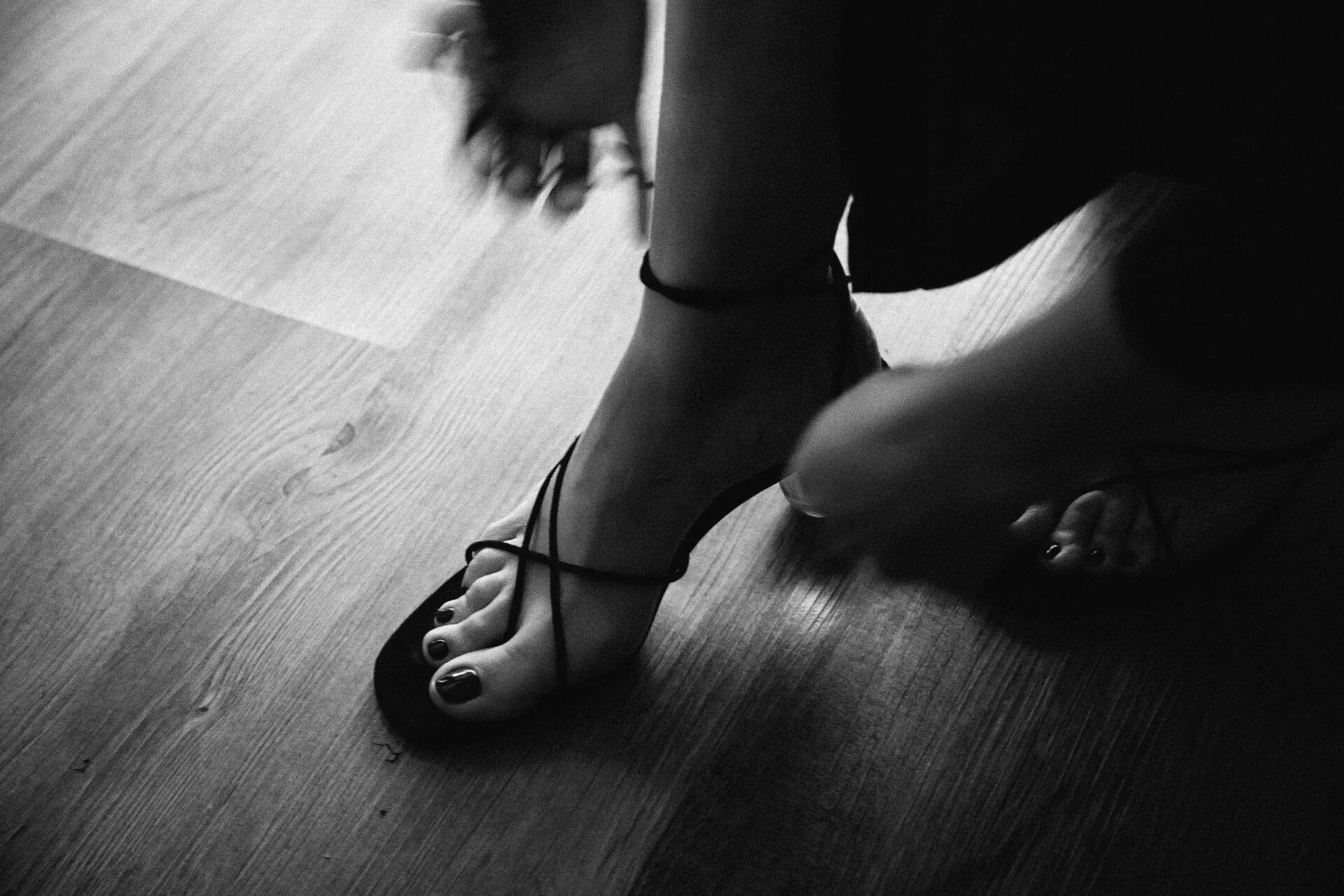 Black and white photo of a woman putting on a strappy high heel shoe on her foot on a wooden floor.