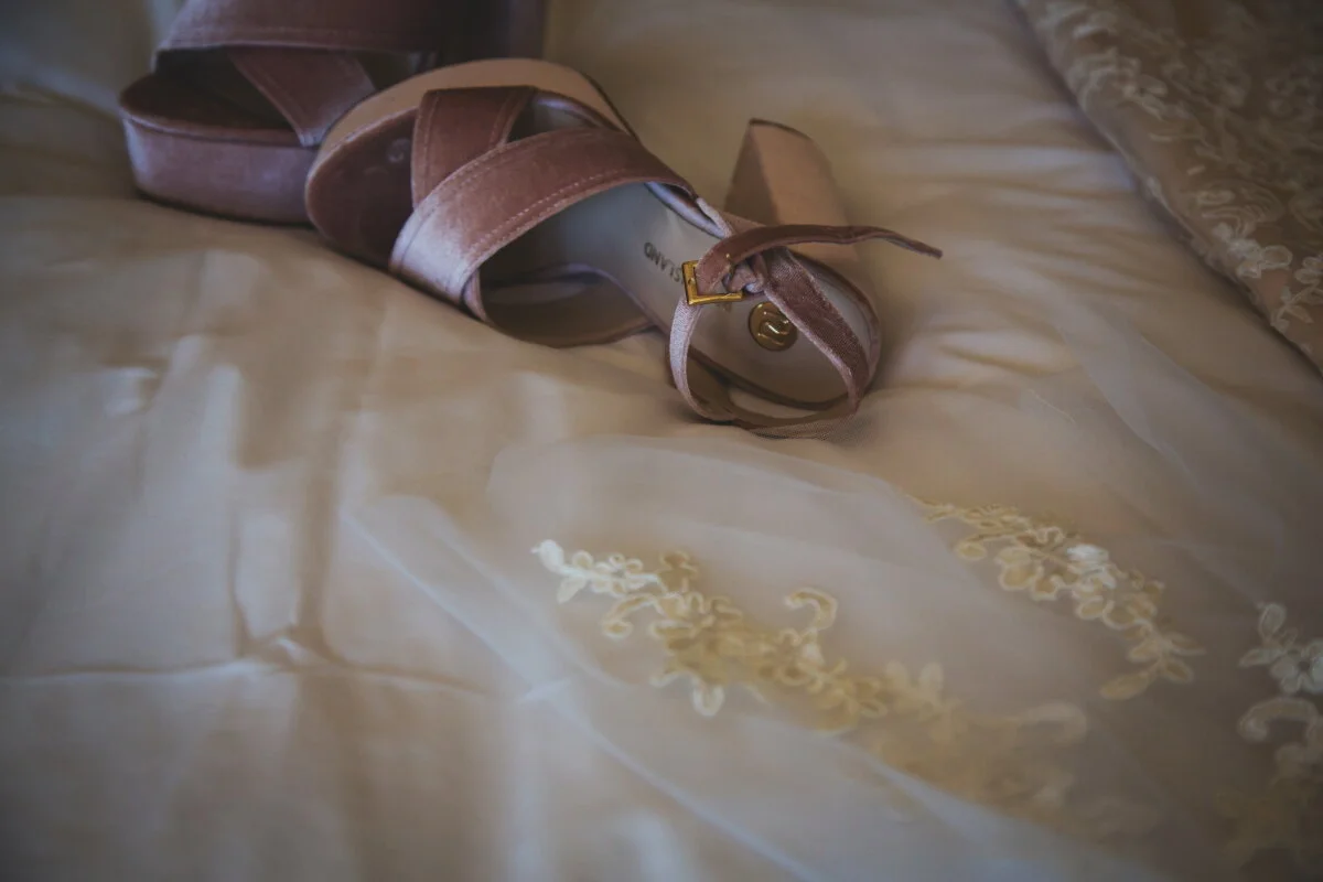 A pair of pink high-heeled shoes with ankle straps resting on a cream-colored bed with embroidered floral patterns.