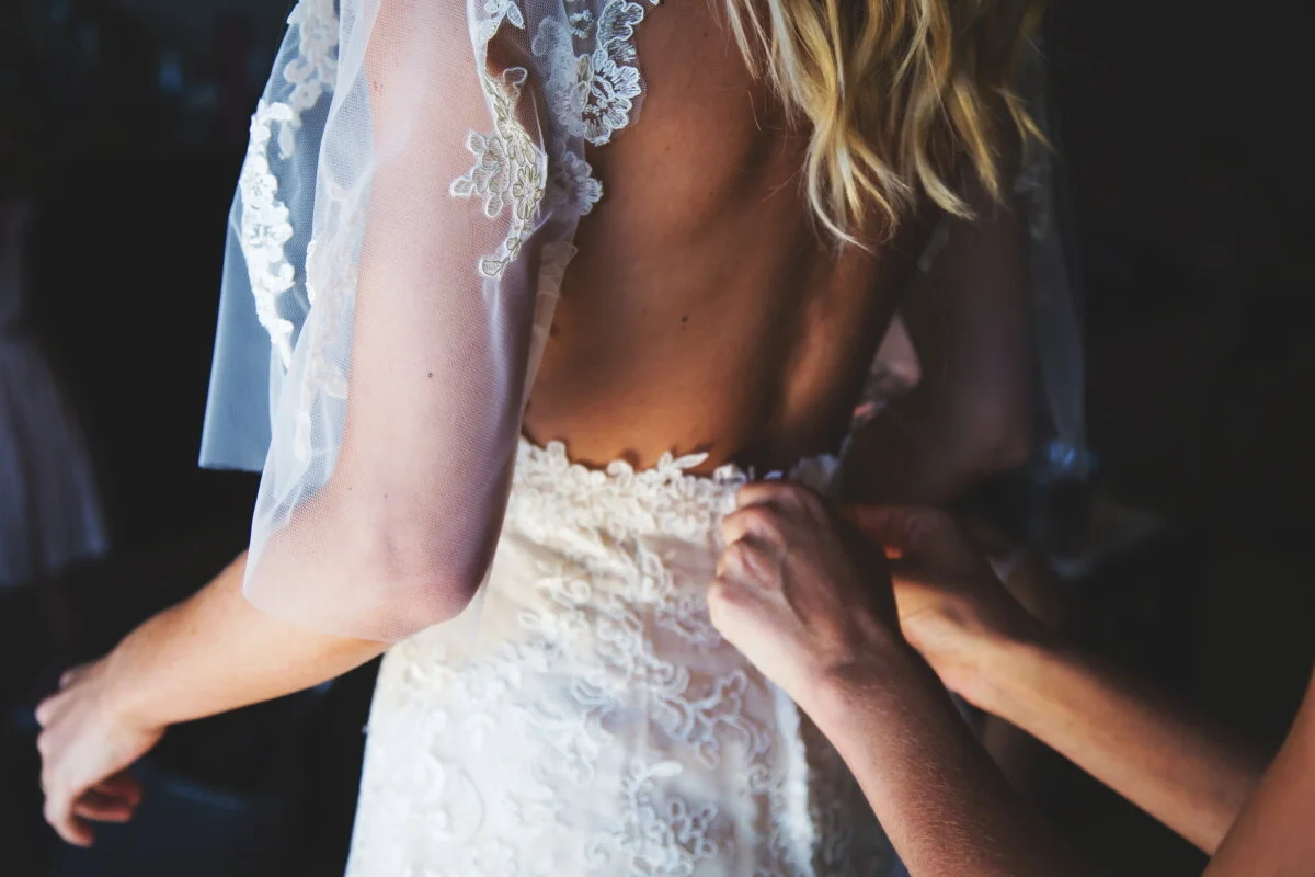 Close-up of a bride in a wedding gown with lace details, as someone helps her get dressed.