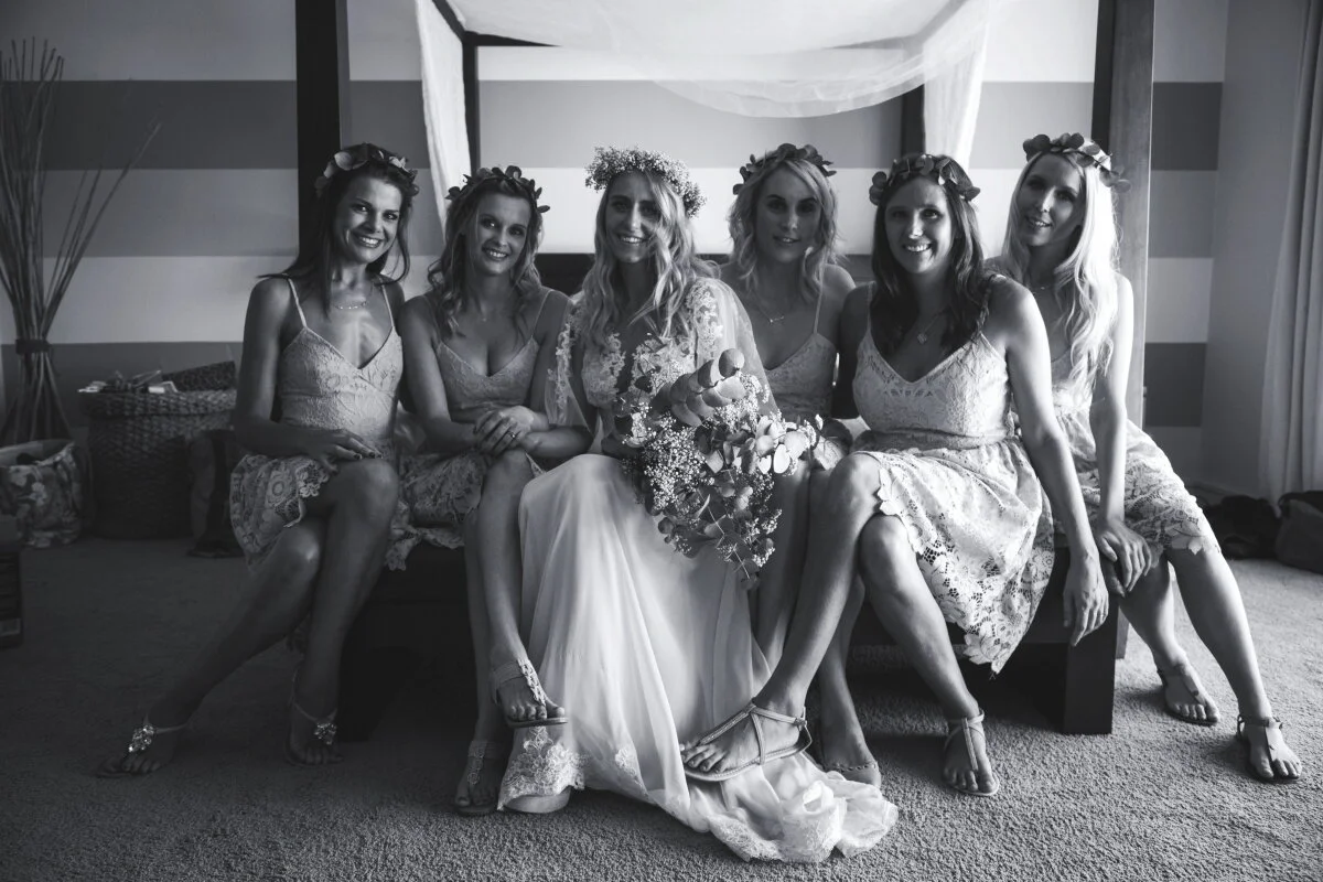 Black and white photo of a bride and five bridesmaids sitting on a bed with a canopy, all smiling, wearing dresses and floral crowns, with the bride holding a bouquet of flowers.