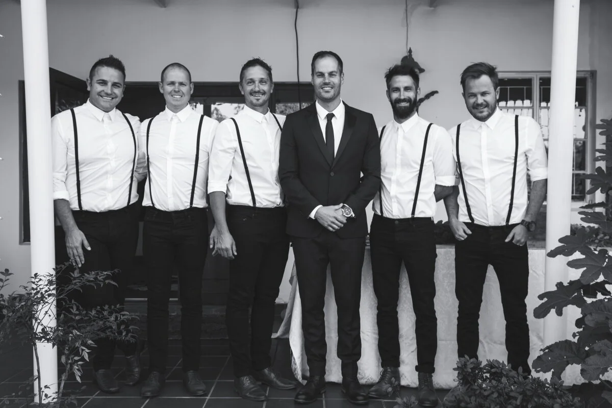 Group of seven men dressed in white shirts with suspenders, one man in a suit, standing together indoors and smiling for the camera.