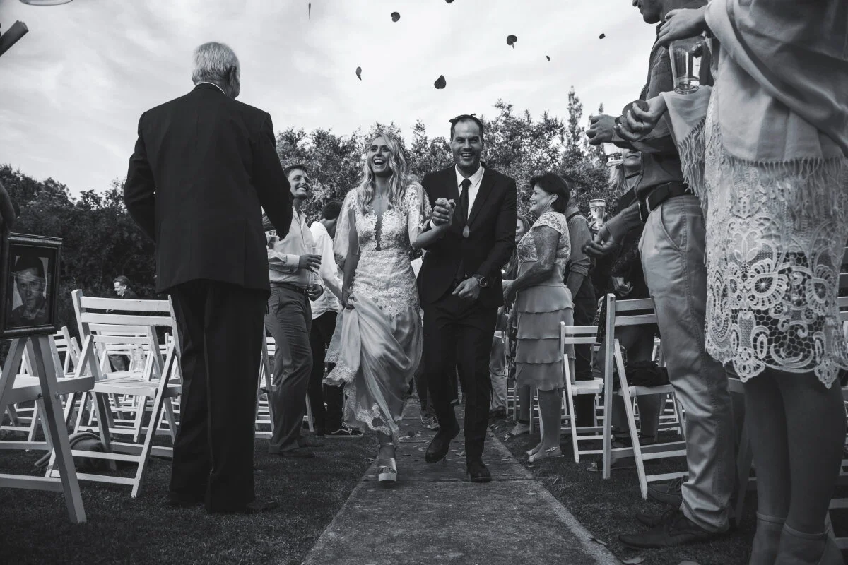 Black and white photo of a wedding ceremony with a bride and groom walking down the aisle together, surrounded by guests holding glasses, outdoors with trees and balloons in the background.