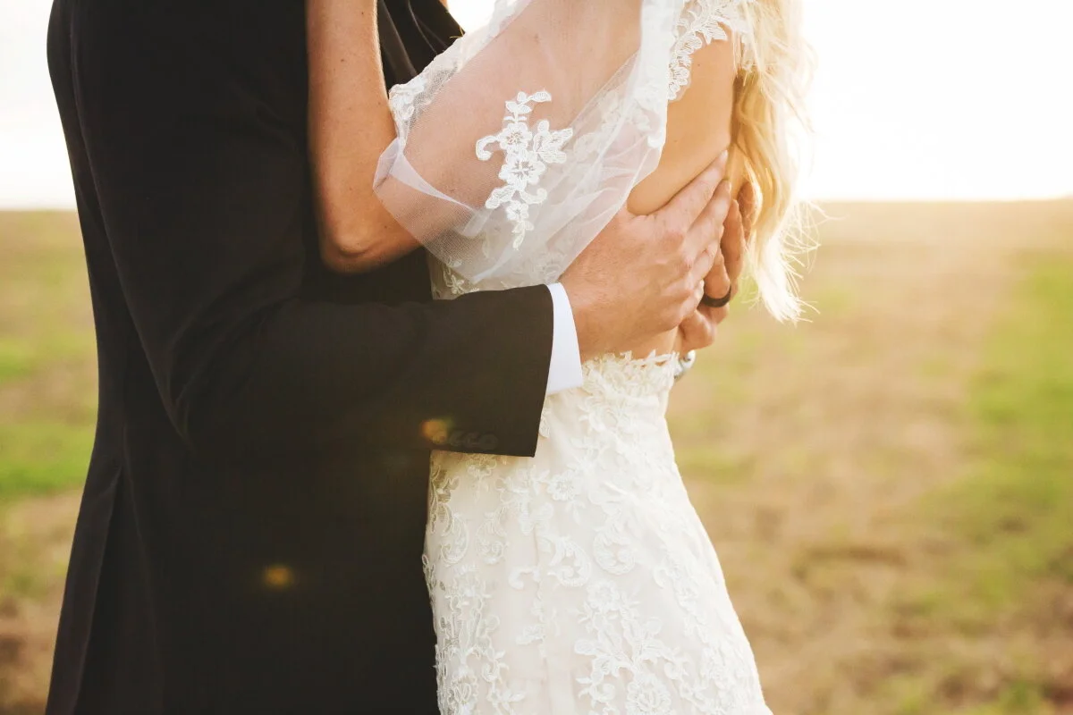A bride and groom embracing outdoors at sunset, with the groom in a black suit and the bride in a white lace wedding dress.