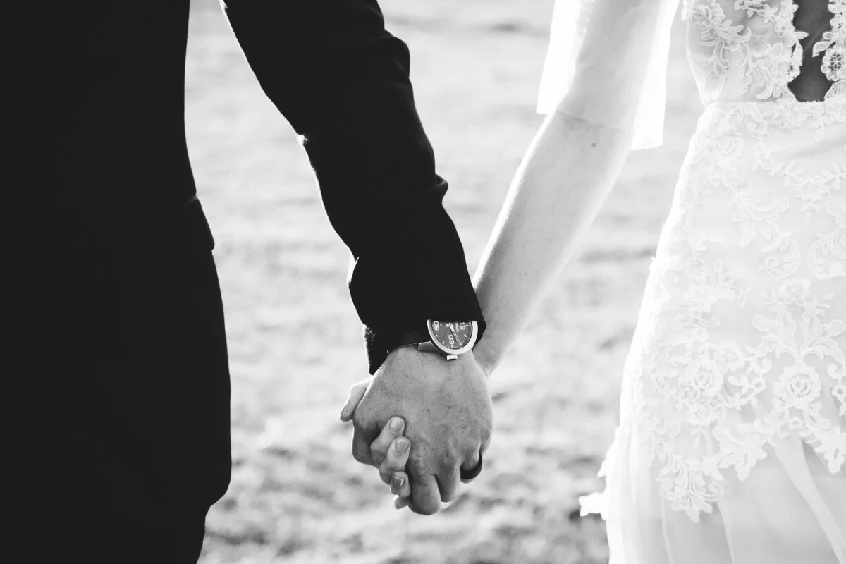 A black and white photo of a couple holding hands, dressed in wedding attire, with the bride in a lace gown and the groom in a suit, on a beach.