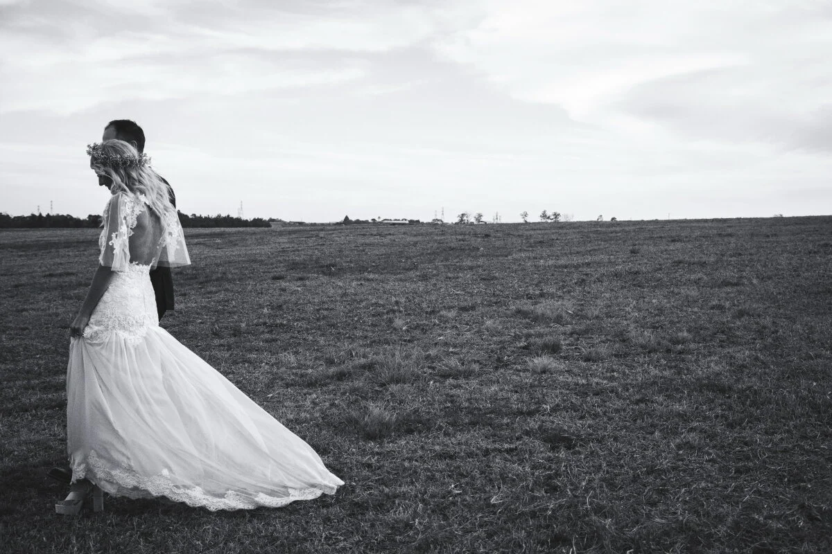 A black and white photo of a bride in a wedding gown standing in a large open field, with a groom standing behind her, both facing downward.