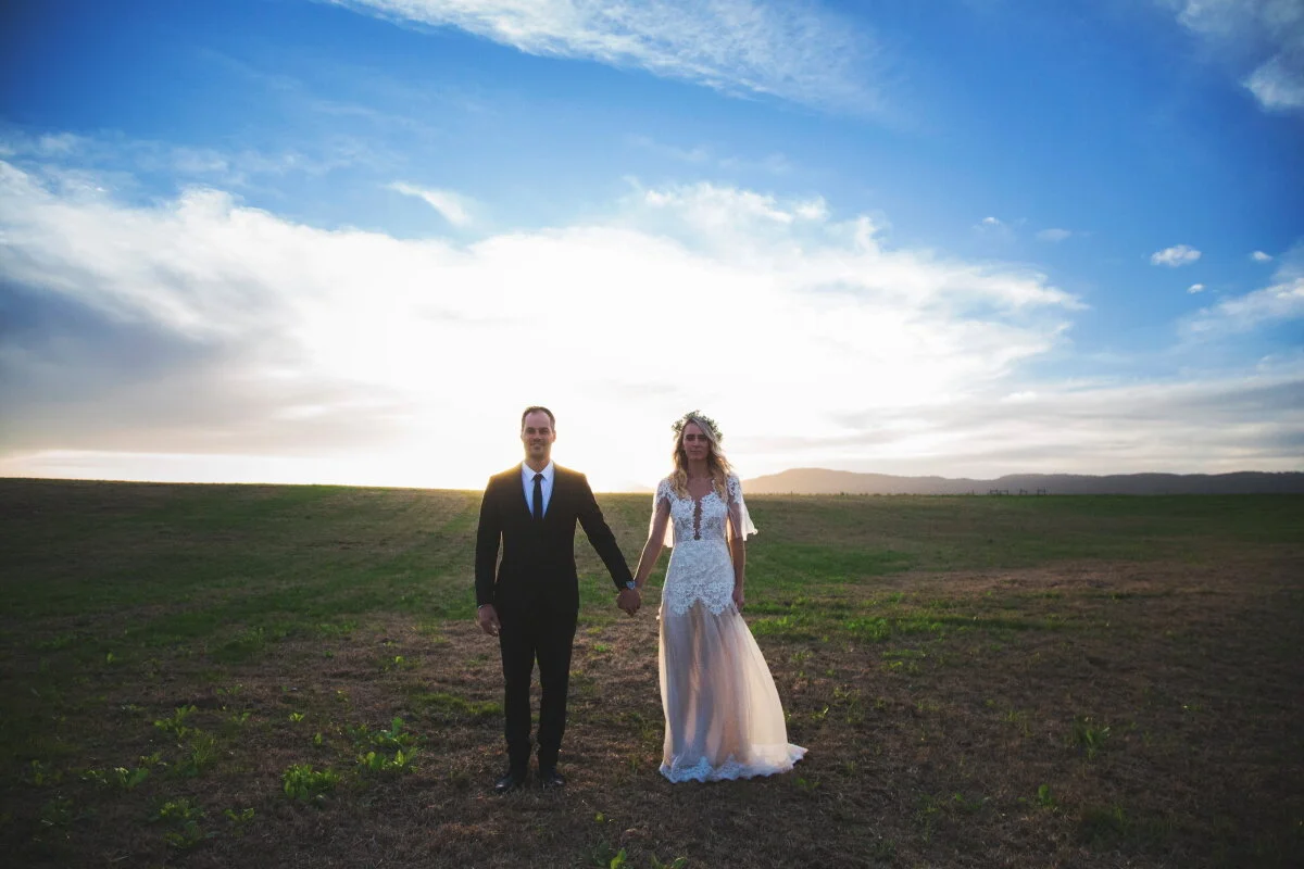 A bride and groom walking hand in hand in an open field during sunset, with a bright sky and clouds in the background.