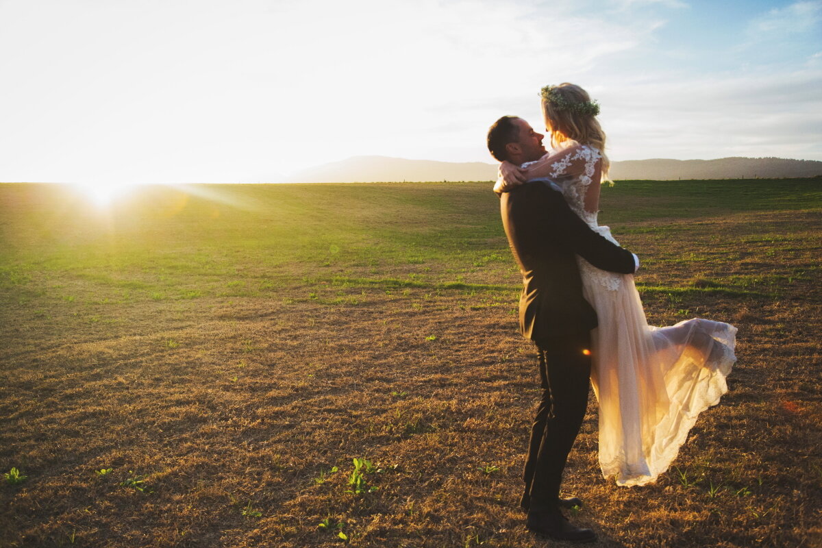 A groom in a black suit carrying a bride in a white wedding dress in a field at sunset.