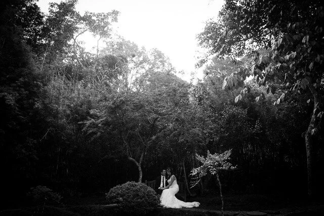 A bride in a white dress sitting on a bench in a lush, wooded outdoor setting.