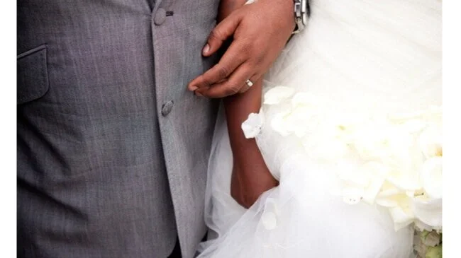 Close-up of a bride and groom holding hands during a wedding, focusing on their clasped hands and wedding attire.