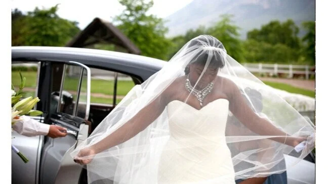 Bridal bride in a white strapless gown and veil getting into a vintage car outdoors.