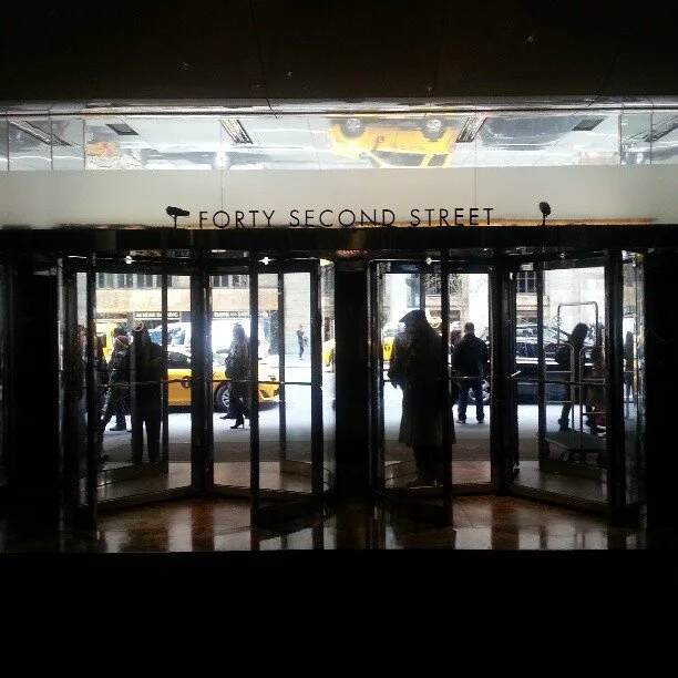 Entrance doors at a building's lobby with a sign reading 'Forty Second Street' above the doors, with people and yellow taxis outside.