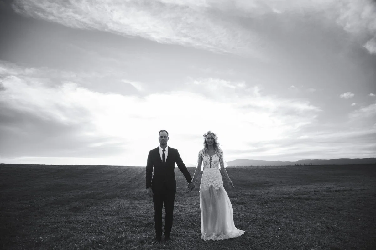 Black and white photo of a bride and groom standing outdoors, holding hands, with a cloudy sky and distant mountains in the background.