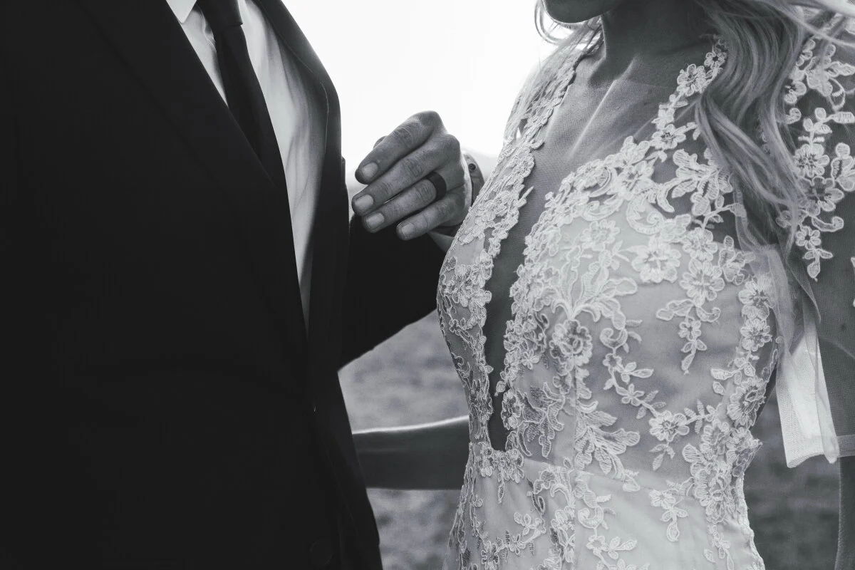 A bride and groom holding each other's hands. The bride is wearing a lace wedding dress and the groom is in a black suit with a white shirt and black tie.