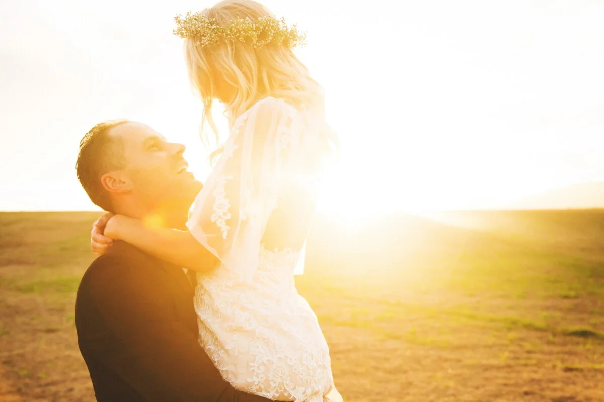 A man and a woman, standing in an open field at sunset. The man is lifting the woman, who is wearing a white dress with lace details and a flower crown, while they look at each other and smile. The warm glow of the setting sun illuminates the scene.