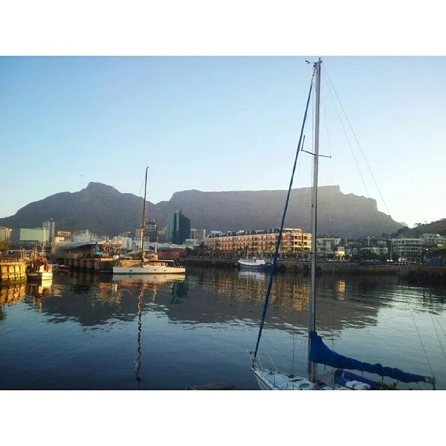 Sailboats docked at a harbor with buildings and Table Mountain in the background.
