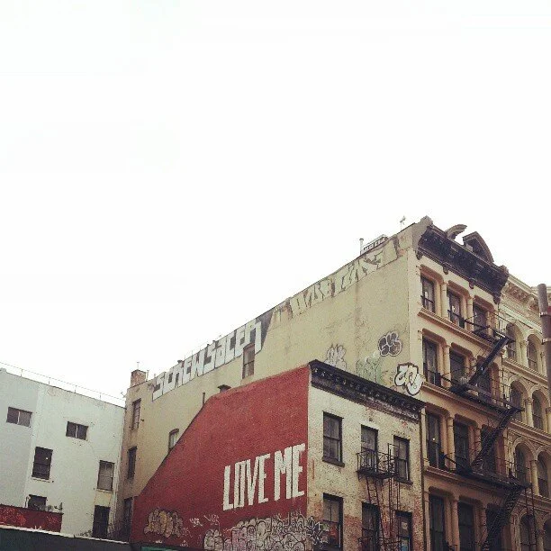 Two buildings with graffiti; one features a large red section with white text saying "LOVE ME", and the other has a beige facade with windows and fire escapes.
