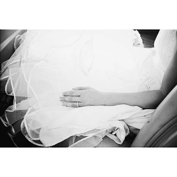 Bride sitting in the back of the car, wearing a wedding ring, with their arm resting on on her lap over the netting, in a black and white photo.