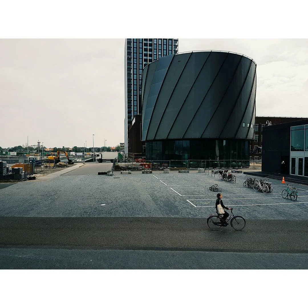 A person riding a bicycle in front of a modern building with a curved, dark exterior. There are bicycles parked nearby and some construction equipment in the background.