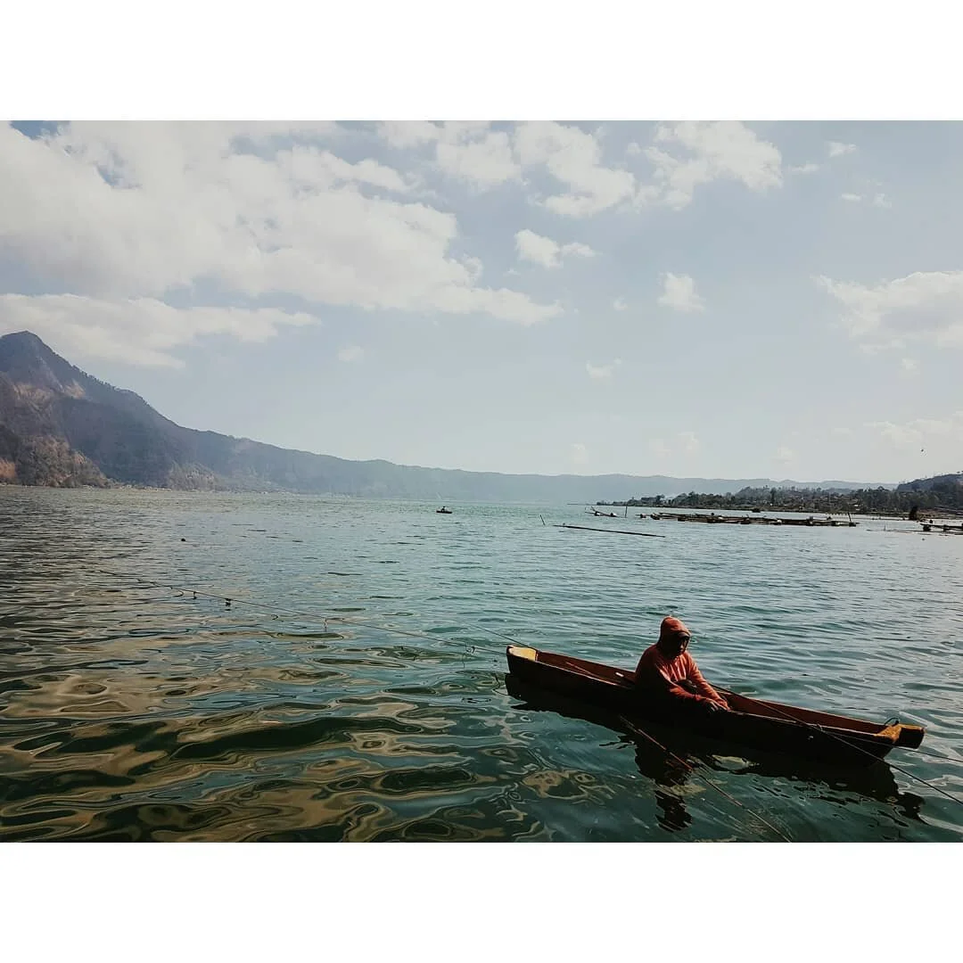 A person in a small wooden boat on a calm lake with mountains and a partly cloudy sky in the background.