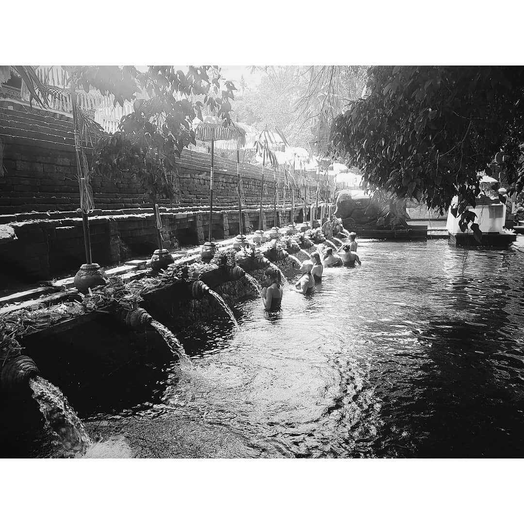 People bathing in a row of small water fountains in a narrow outdoor temple pool, with trees and plants overhead, in black and white.