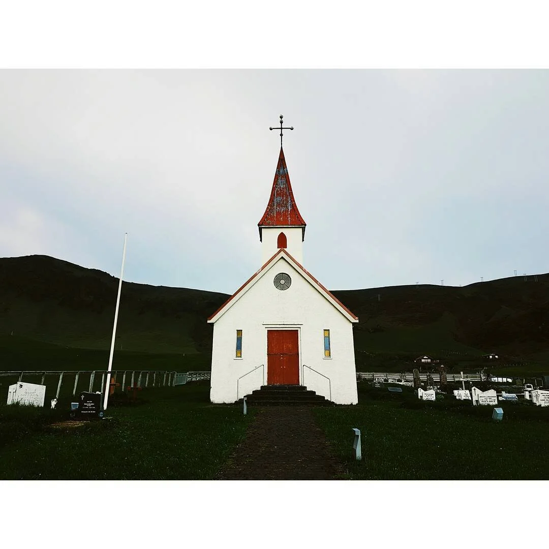 Small white Icelandic church with a red roof and door, surrounded by green grass and grave markers, against a backdrop of rolling hills under a cloudy sky.