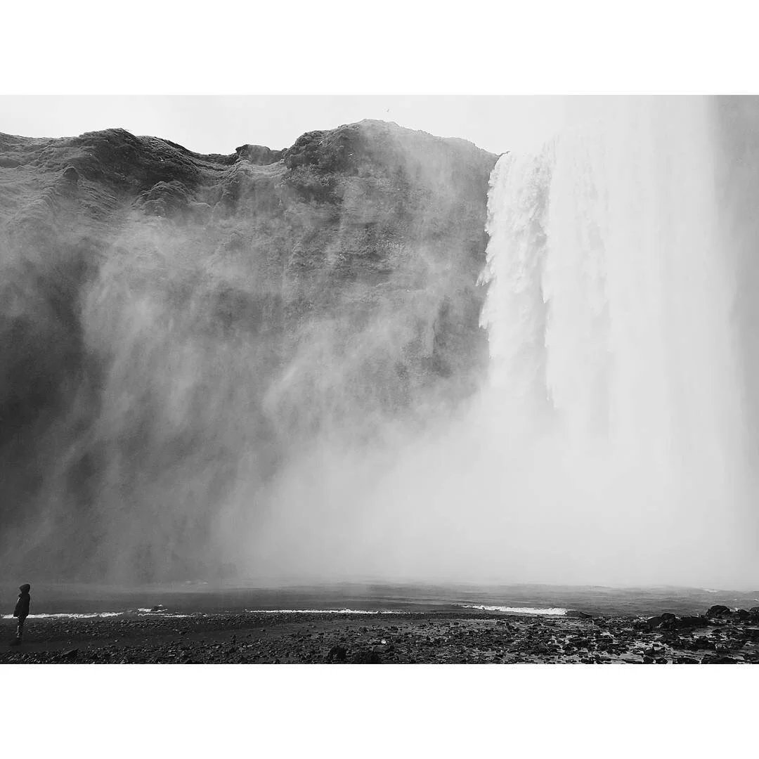 Black and white photograph of a large Icelandic waterfall with mist rising from it, and a solitary person standing at the bottom observing the scene.