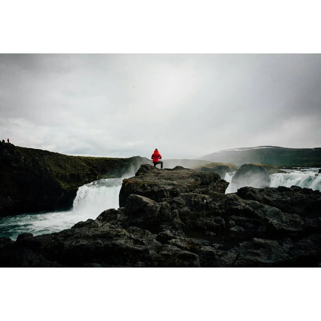 A person in a red jacket sitting on rocks near a waterfall in a cloudy, mountainous landscape.