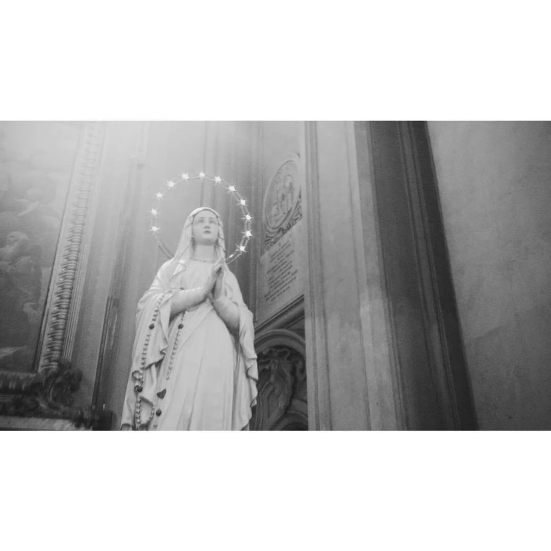 Black and white photo of a religious statue of the Virgin Mary with a halo, holding prayer beads with hands in prayer, in a church interior.