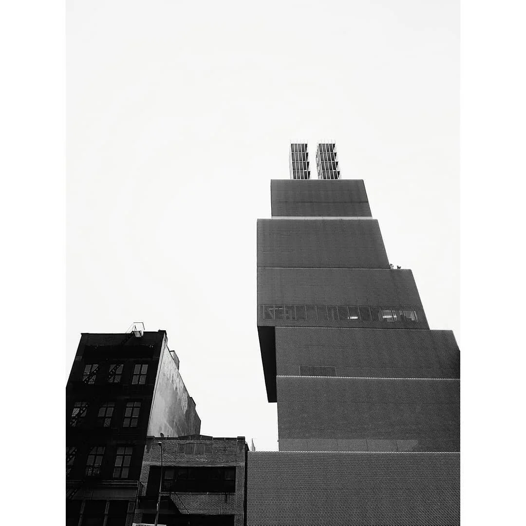 Black and white photo of tall modern New York skyscraper with stacked, offset box-like sections against a plain sky, surrounded by older buildings.