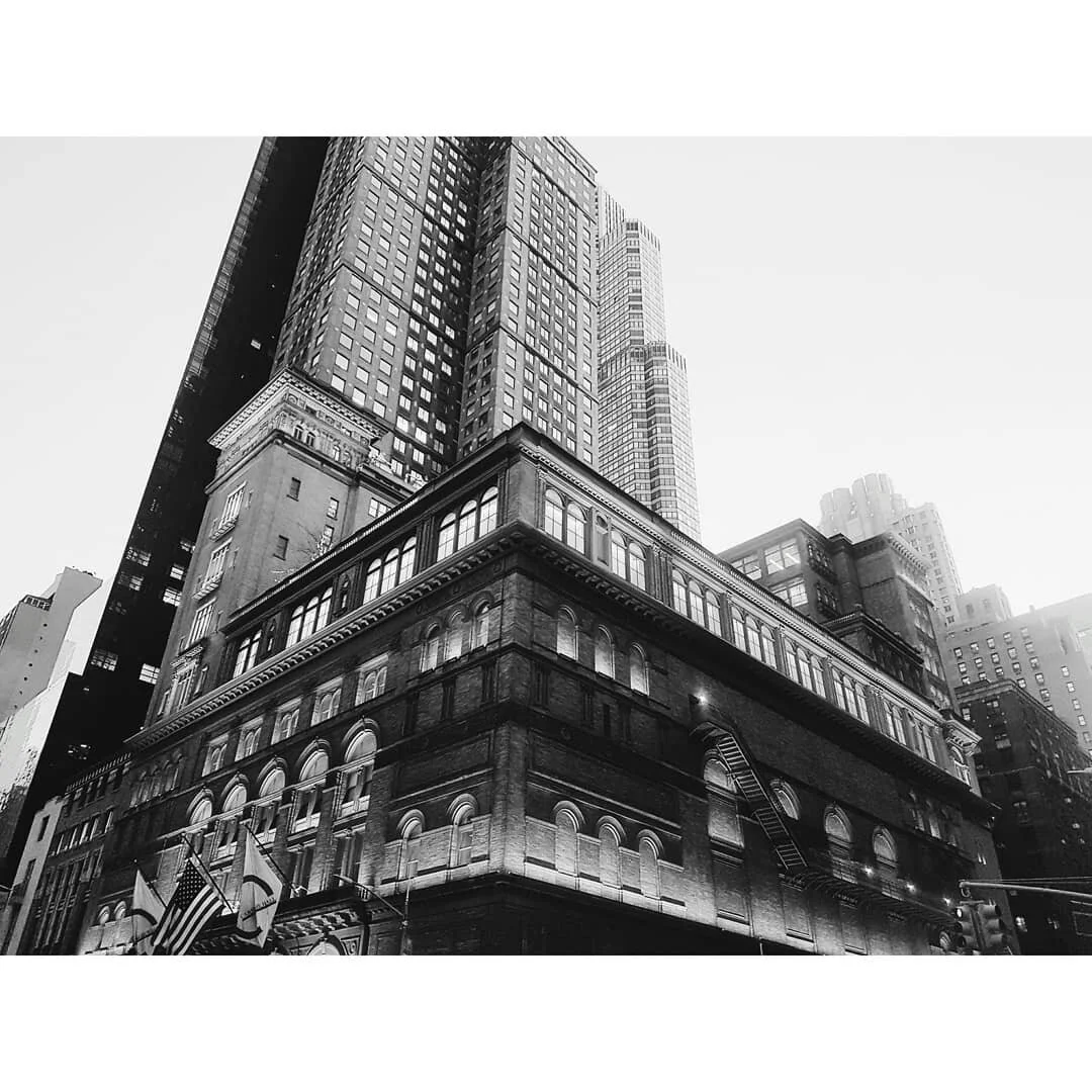 Black and white photo of tall skyscrapers in an urban cityscape, featuring a historic multi-story building in the foreground with arched windows and detailed architectural elements.