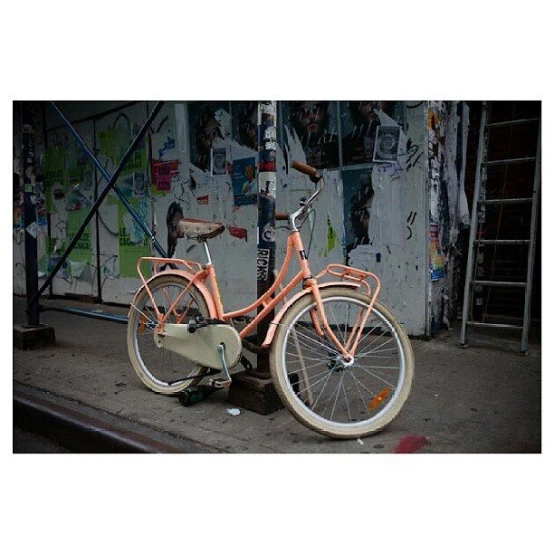 A pink vintage-style bicycle with white-tread tires is parked on the sidewalk in front of a graffitied wall covered with posters and stickers.