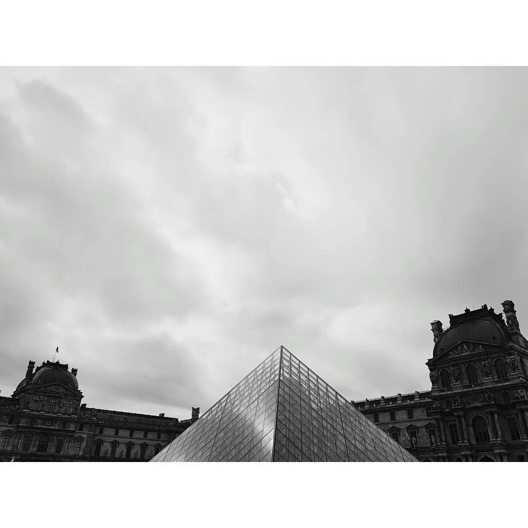 Black and white photo of the Louvre Museum in Paris with a modern glass pyramid in the foreground and historical buildings in the background, under a cloudy sky.