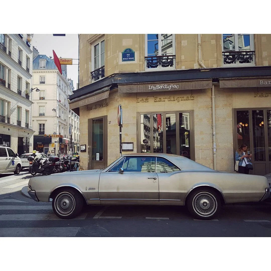 A vintage silver Cadillac parked on a city street in front of a French restaurant called 'Les Sardignac' on Rue Boule Rouge in Paris. A woman is standing near the restaurant entrance, and there are other cars and motorcycles on the street.