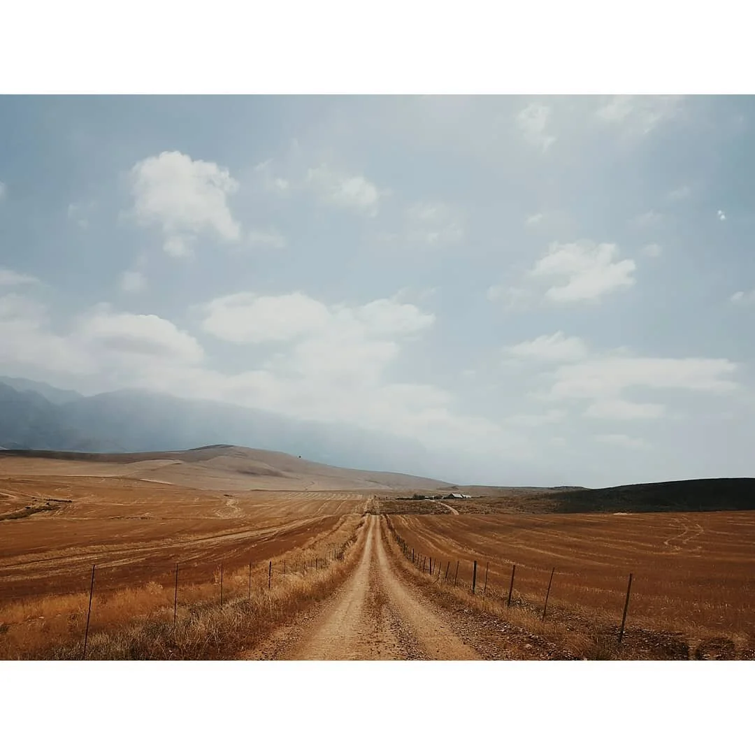 A dirt road running through a vast, open plain with dry grass, flanked by fencing on both sides, leading toward distant hills under a partly cloudy sky.