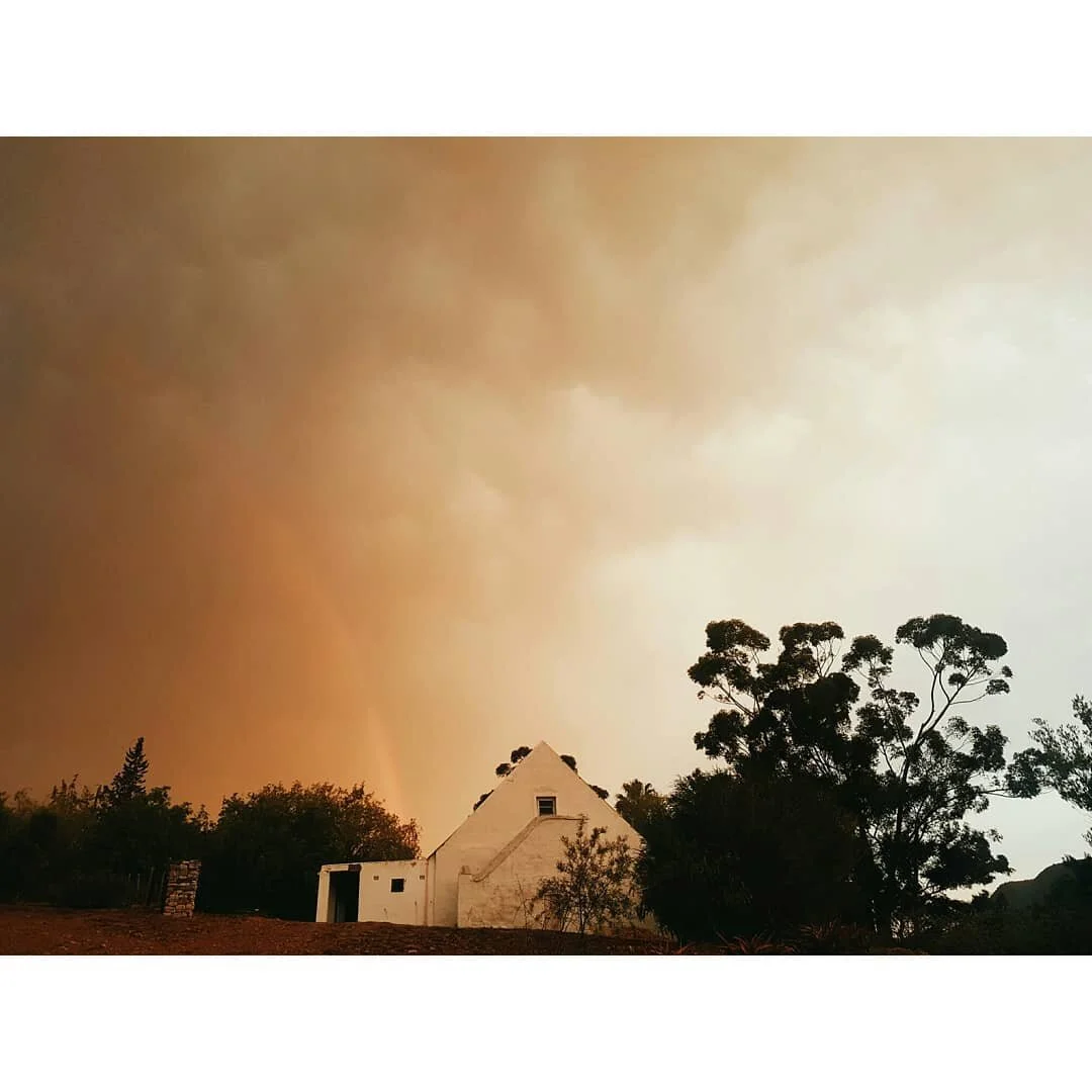 A small white Karoo building with a triangular roof, surrounded by trees, under an ominous orange sky.