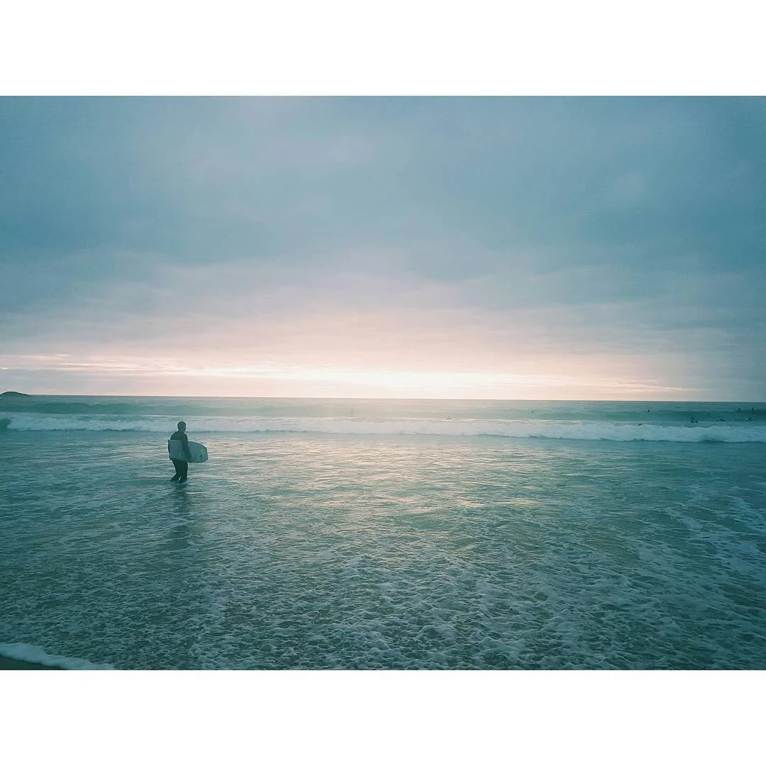 A person holding a surfboard standing in the shallow water at the beach during sunset or sunrise.