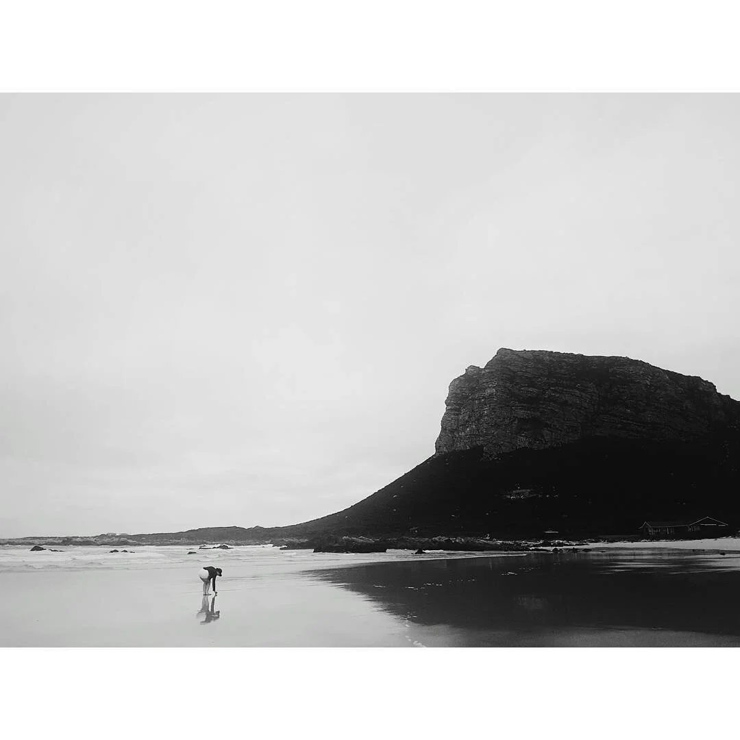 A black and white photo of a person walking on a beach with a large cliff in the background and the ocean waves in the distance.